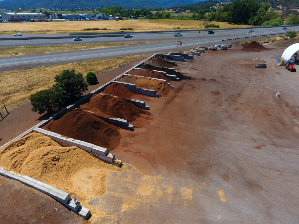 Aerial view of a landscaping supply yard with organized piles of mulch, soil, and bark along a highway.