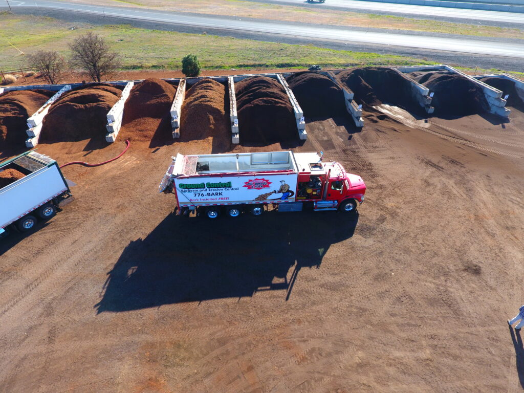 Aerial view of a red bark blower truck parked at a mulch yard, with large piles of bark and compost materials stored in divided concrete bays beside a roadway.