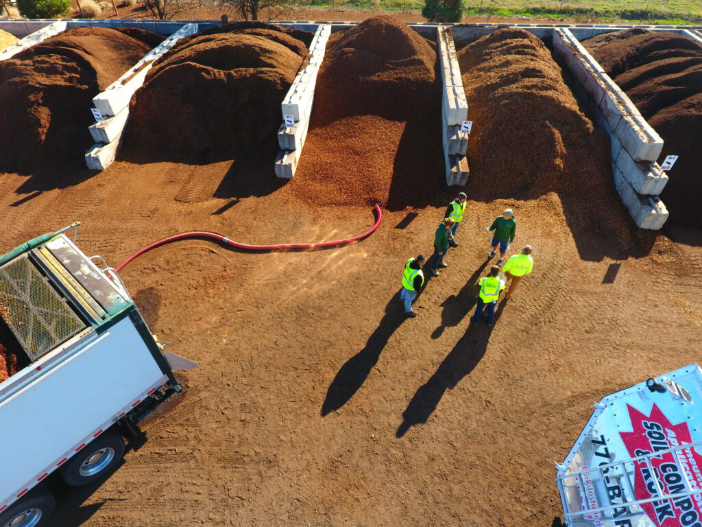 Aerial view of a mulch yard with large, separated piles of brown mulch, a blower truck with a red hose, and several workers in high-visibility safety vests standing and talking on the dirt lot.