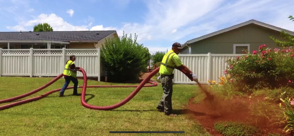 Two workers in high-visibility shirts use a large blower hose to spread red bark mulch along a flower bed beside a white vinyl fence in a residential backyard on a sunny day.