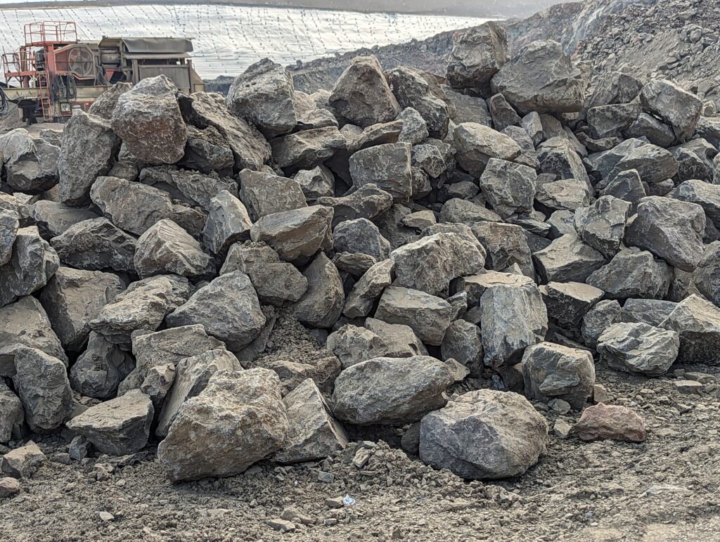 Pile of large, rough-cut gray natural stone boulders stacked at a quarry site, with heavy equipment and terraced rock walls visible in the background.
